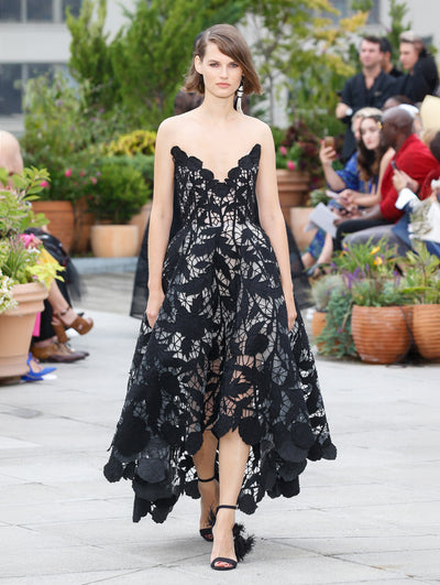 The model walks the runway in a black strapless semi-sheer cocktail dress with a threadwork cutout leaf design. Behind her, the blurred background reveals the audience and potted plants.
