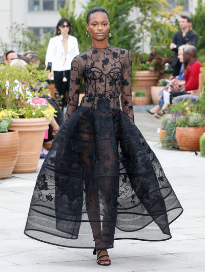 The model walks the runway in a black long-sleeve semi-sheer  evening gown with a bustier bodice and threadwork embroidered leaves. Behind her, the blurred background reveals the audience and potted plants.