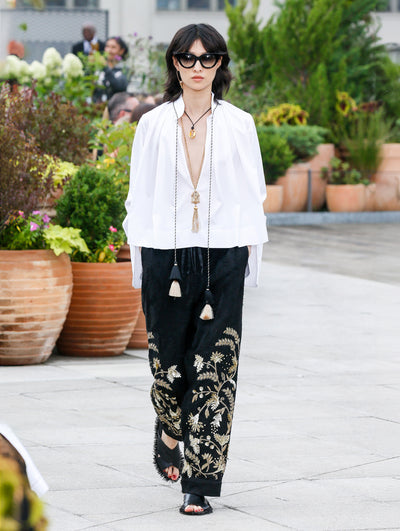 The model walks the runway in a white long-sleeve tie-neck blouse and black velvet trousers with gold threadwork embroidered florals. Behind her, the blurred background reveals the audience and potted plants.