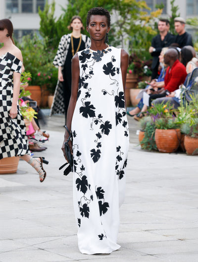 Holding a black clutch, the model walks the runway in a white sleeveless column evening gown printed with black leaves. Behind her, the blurred background reveals the audience and potted plants.