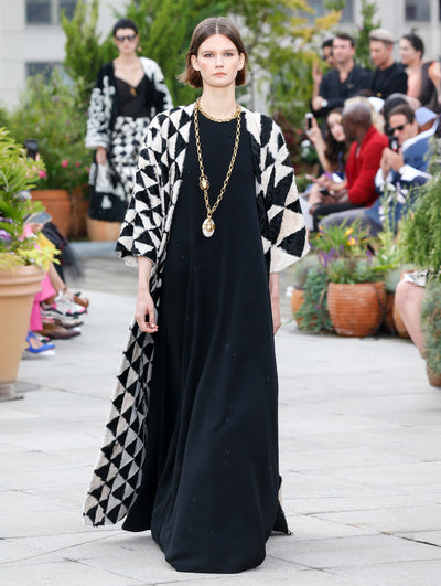 The model walks the runway in a short-sleeve floor-length coat with a black and white geometric pattern layered over a black caftan. Behind her, the blurred background reveals the audience and potted plants.