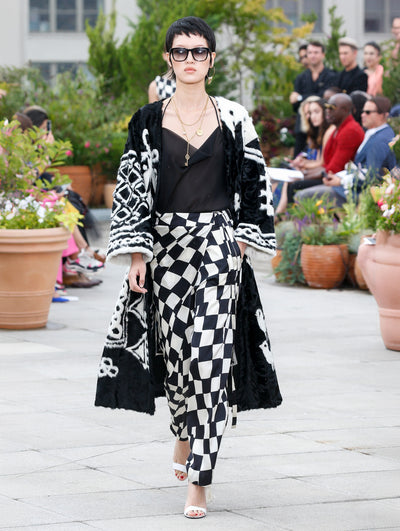 Wearing black sunglasses, the model walks the runway in a black and white fur coat with a geometric design, layered over a black blouse and a silk skirt with a black and white checkered design. Behind her, the blurred background reveals the audience and potted plants.
