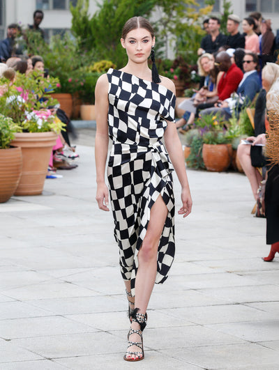 The model walks the runway in an off-shoulder silk evening gown with a black and white checkered design. Behind her, the blurred background reveals the audience and potted plants.