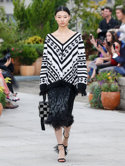 Holding a black and silver checkered cube-like handbag, the model walks the runway in a long-sleeve V-neck knit pullover with a black and white geometric design and fringe trim, paired with a sequin embroidered black skirt with feather trim. Behind her, the blurred background reveals the audience and potted plants.