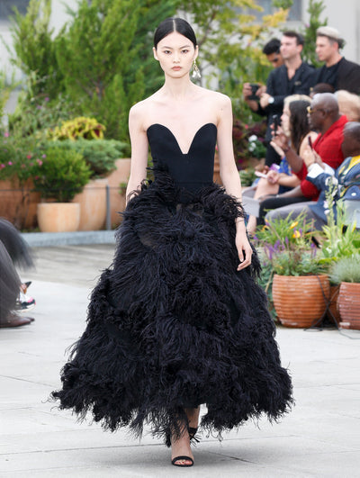 The model walks the runway in a black evening gown with a strapless sweetheart wool bodice and a feather skirt. Behind her, the blurred background reveals the audience and potted plants.