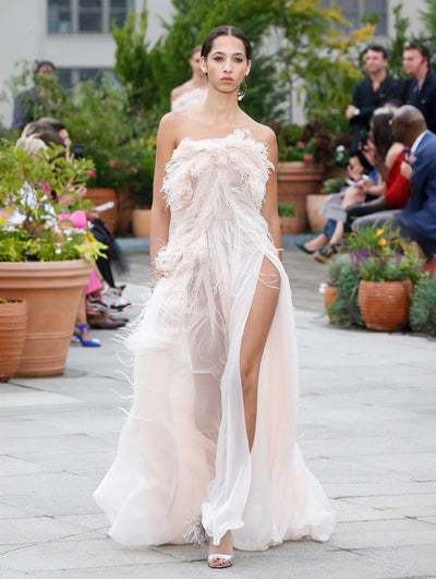 The model walks the runway in an ivory strapless evening gown with feather embroidery concentrated on the bodice that fades to a delicate scatter on the skirt. Behind her, the blurred background reveals the audience and potted plants.