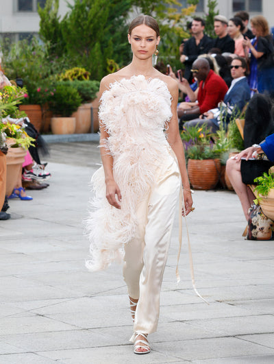The model walks the runway in an ivory strapless feather blouse with an asymmetrical hemline, paired with ivory satin trousers. Behind her, the blurred background reveals the audience and potted plants.