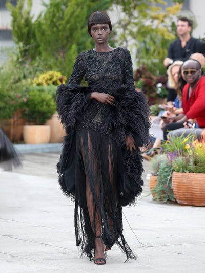 The model walks the runway in a black feather shawl, layered over a black long-sleeve threadwork embroidered evening gown with a fringe floor-length skirt. Behind her, the blurred background reveals the audience and potted plants.