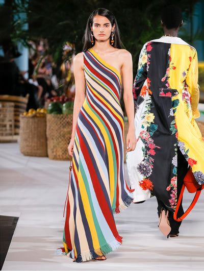 A model wears a rainbow striped one shoulder gown with fringe at the hem