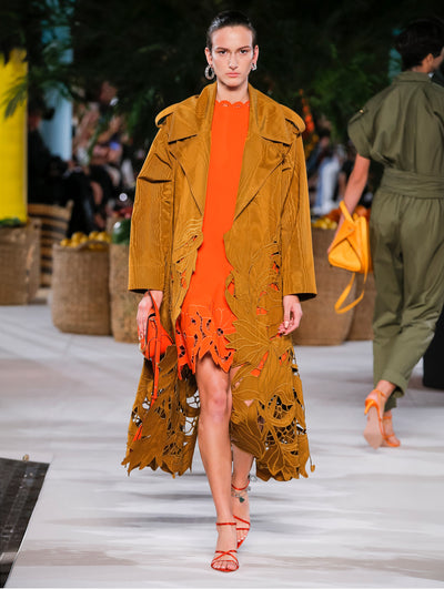 A model wears an orange dress and bronze coat both have floral cut outs at the bottom