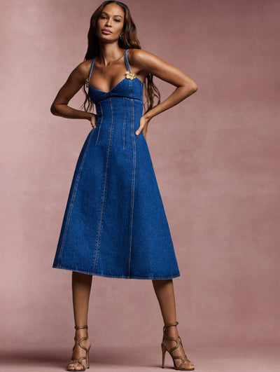 The model wears a denim sleeveless midi dress with a gold flower on each strap from the Spring 2024 collection.